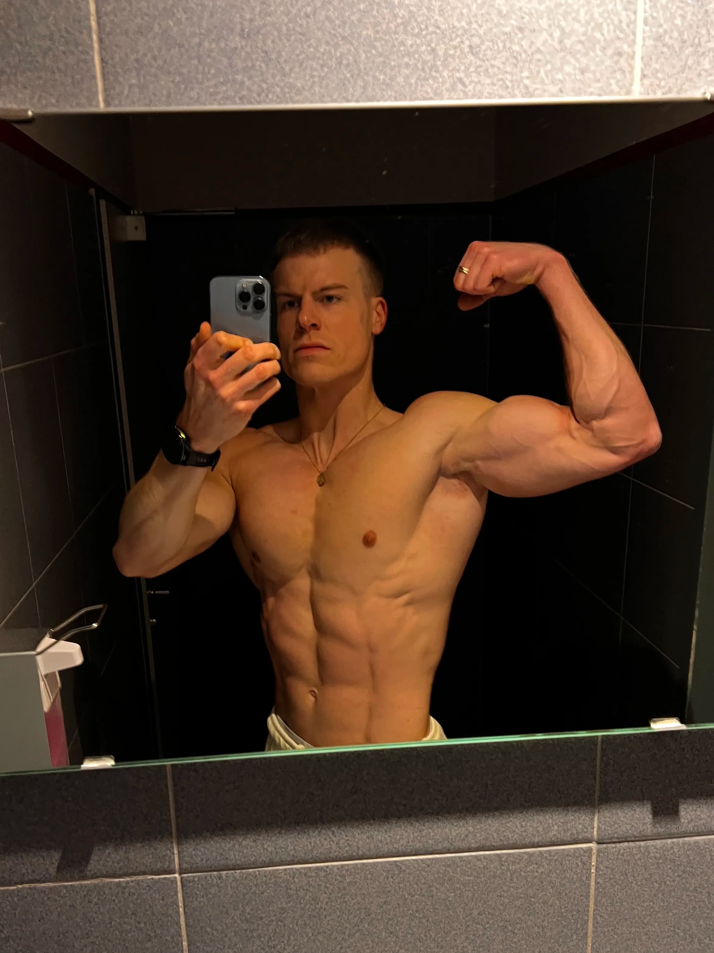 A shirtless man takes a selfie in the mirror showing his muscular physique, flexing his right arm.