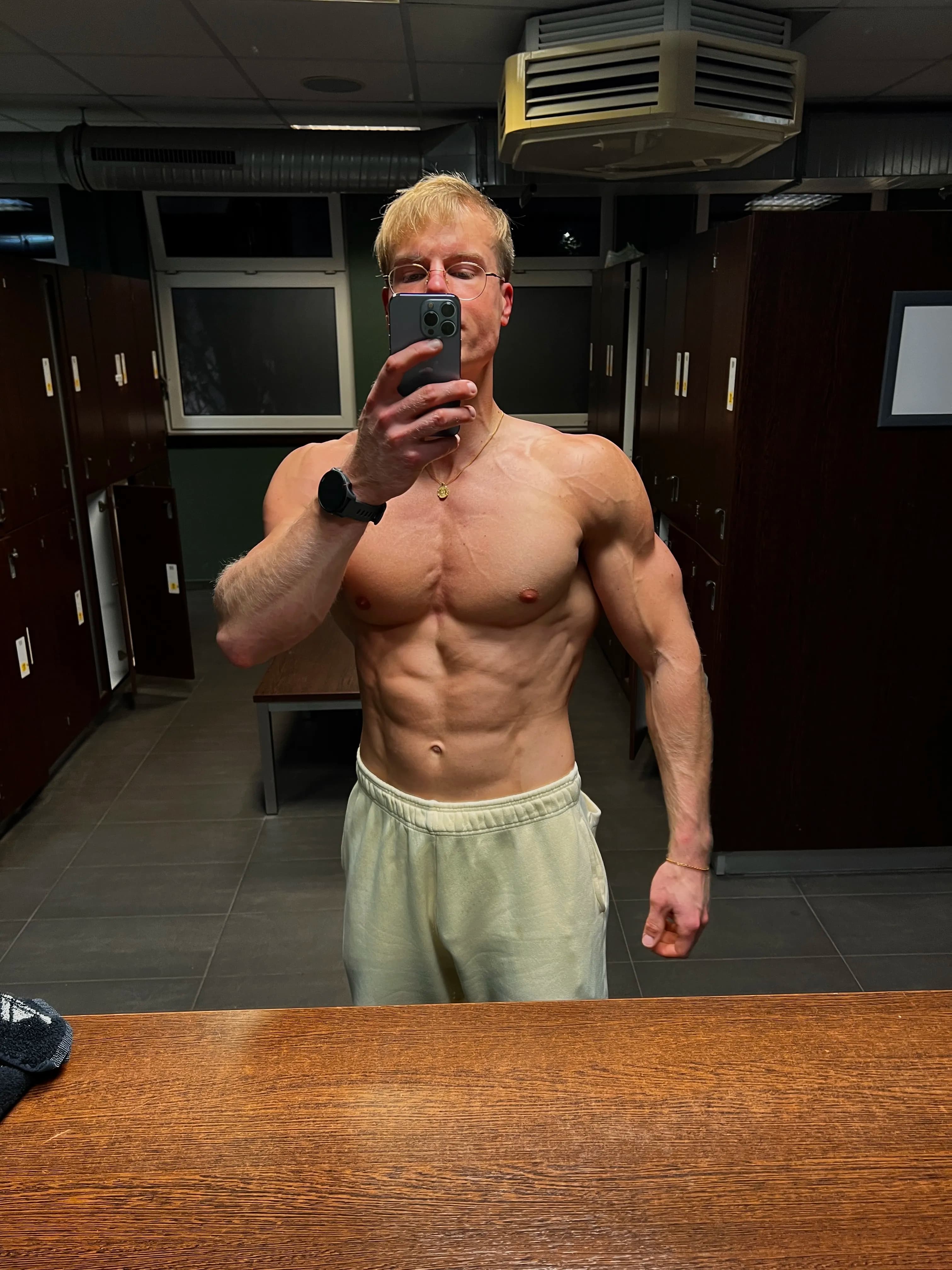 A person taking a mirror selfie in a gym locker room, showing a muscular physique, wearing glasses and light-colored pants.