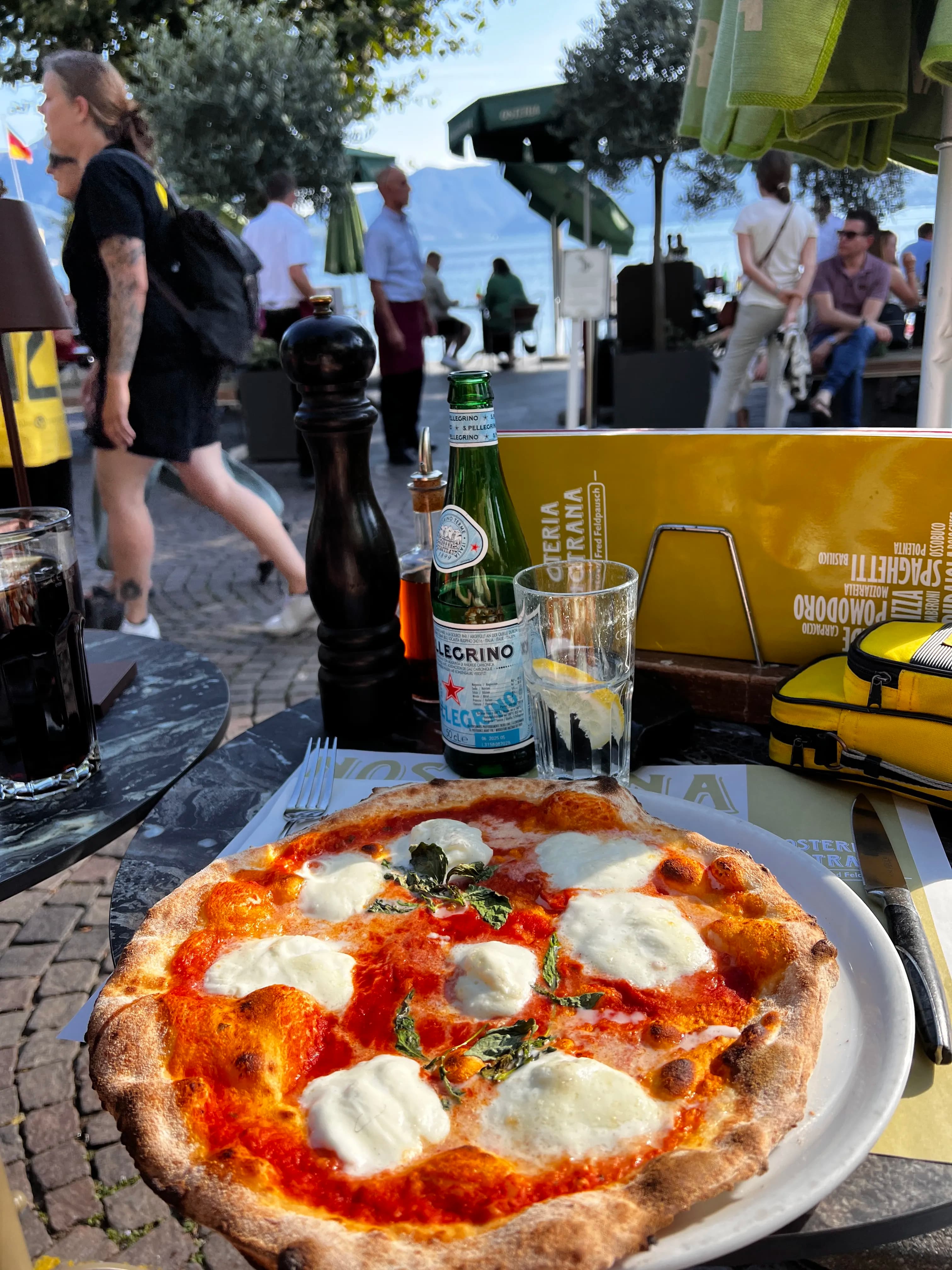 A Margherita pizza with fresh mozzarella and basil sits on an outdoor restaurant table. A bottle of sparkling water and a busy street are in the background.